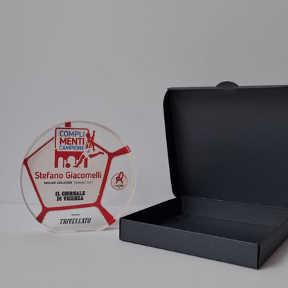 Trofeo rotondo in plexiglass con grafica a pallone da calcio rossa stampata, accanto alla scatola regalo nera